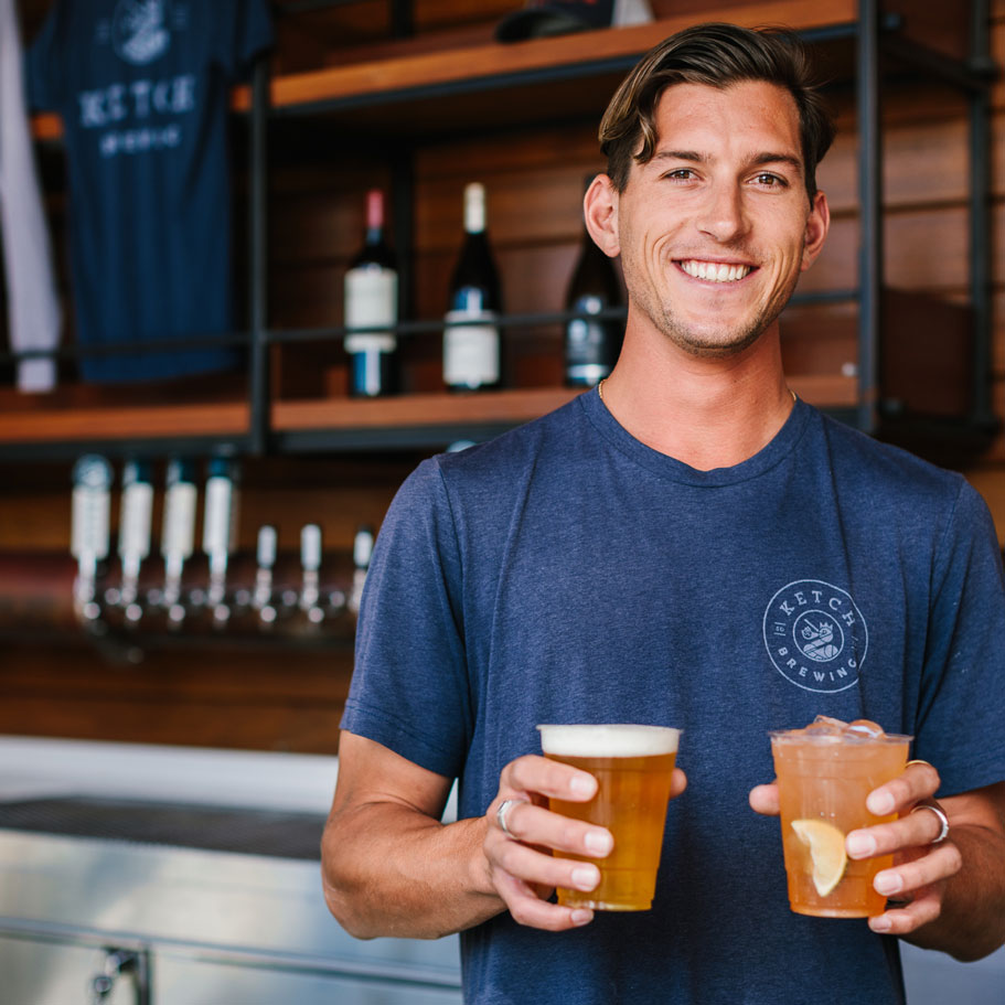 man behind bar holding two cups of ketch beer